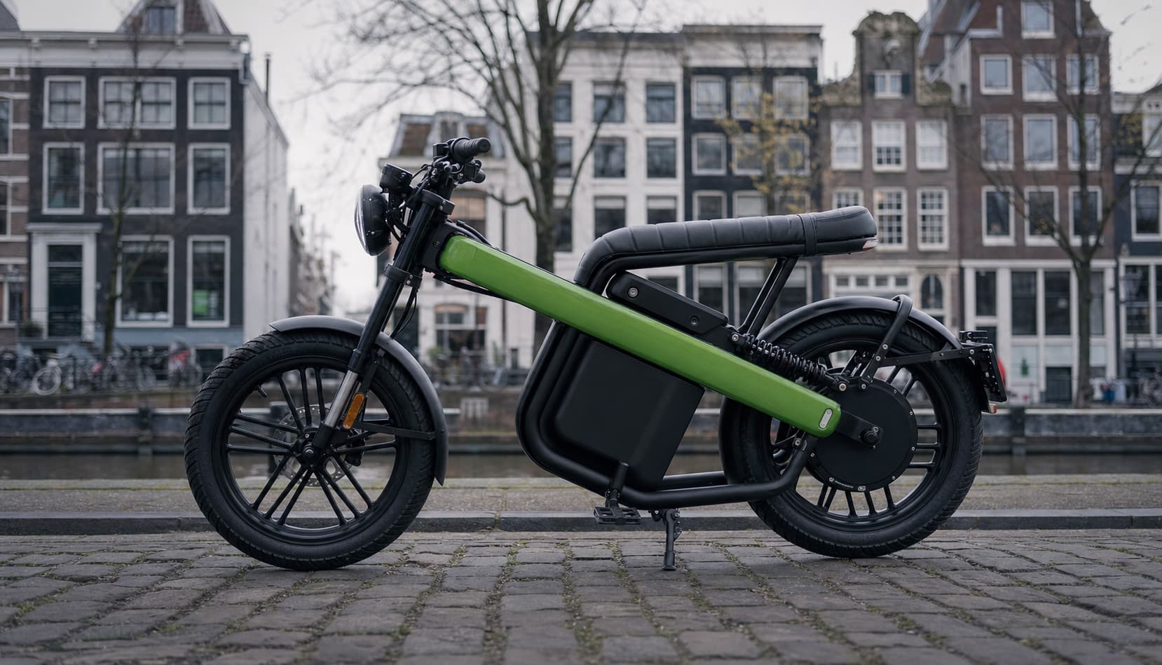 Une moto électrique Brekr Model B au cadre ouvert et aux couleurs vives est garée sur une rue pavée sous une lumière douce de journée nuageuse.