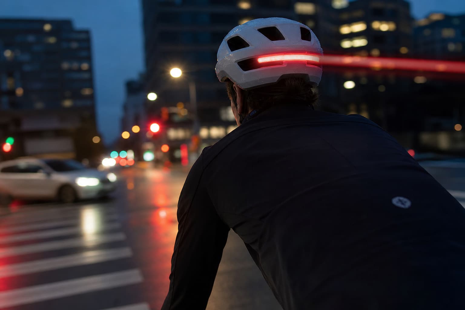 Cycliste portant un casque blanc avec barre LED rouge lumineuse laissant une traînée lumineuse, roulant à un carrefour urbain au crépuscule avec lampadaires et feux de circulation flous en arrière-plan.